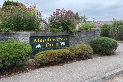 A stone wall with flowering bushes displays a green sign reading "Meadowcrest Farm Estates" with white horse and cow icons. A sidewalk runs in front.