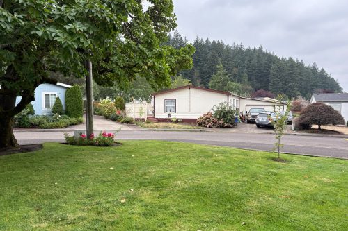 A suburban street with manufactured homes, green lawns, a parked car in a driveway, and trees in the background under a cloudy sky.