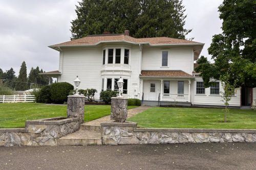 Two-story white house with bay windows and a wraparound porch, surrounded by a manicured lawn and stone fence, on a cloudy day.